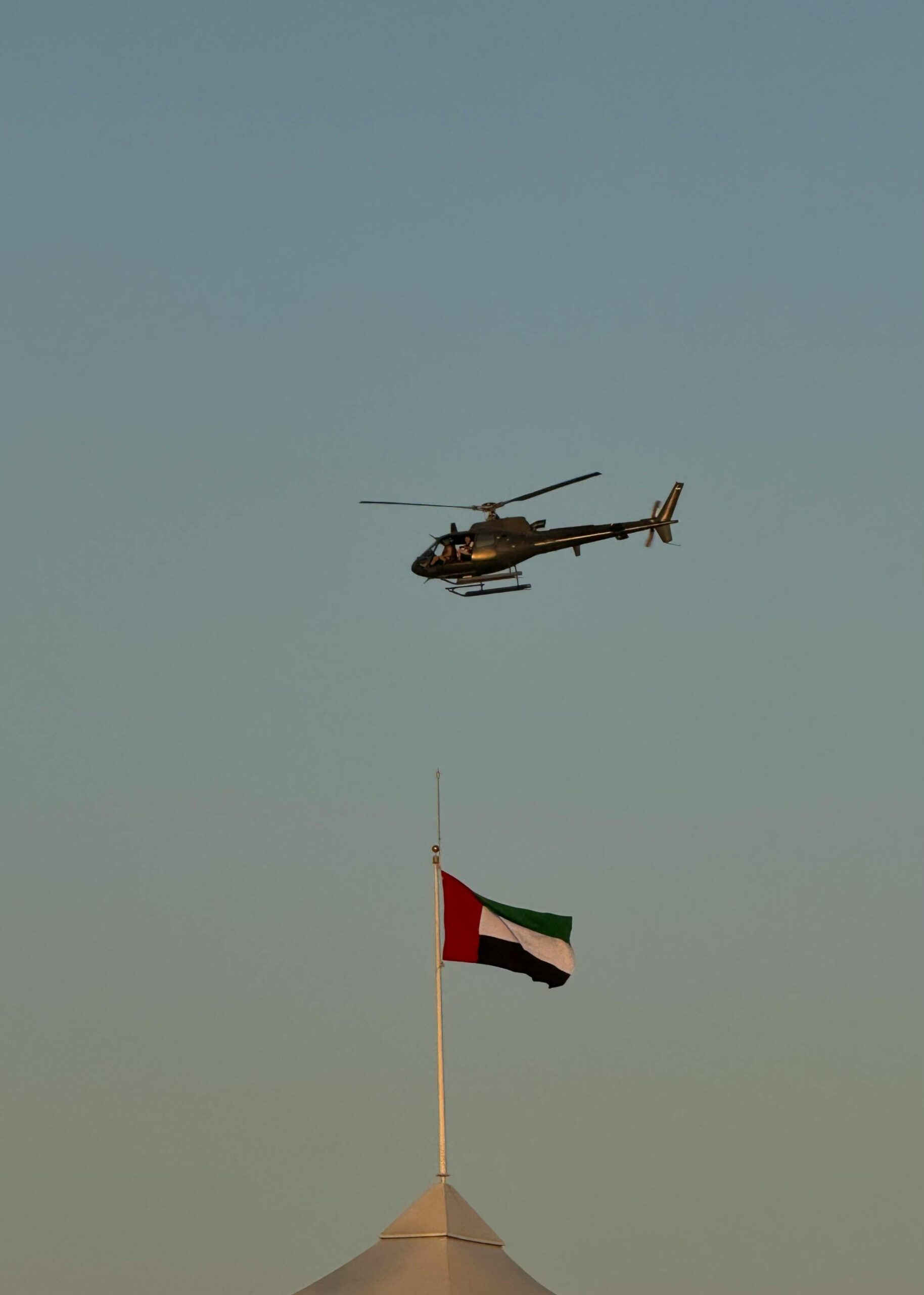A military helicopter flies above the UAE flagpole against a clear sky.